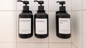 Τρία μαύρα μπουκάλια με μαλακτικό μαλλιών, σαμπουάν και αφρόλουτρο, τοποθετημένα σε έναν τοίχο με πλακάκια.