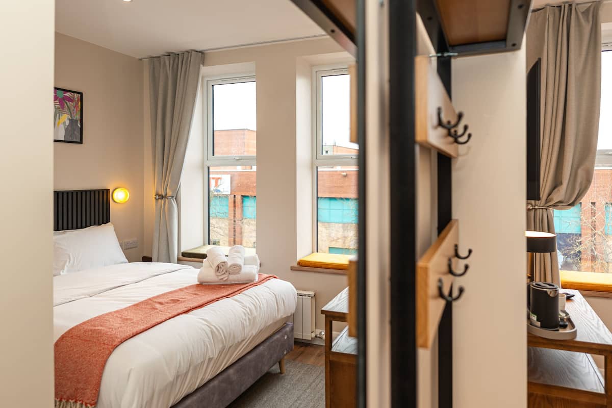 A hotel room with a bed, a desk, a chair, a window with curtains, a radiator, and a wall-mounted hook.