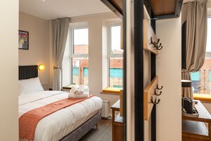 A hotel room with a bed, a desk, a chair, a window with curtains, a radiator, and a wall-mounted hook.