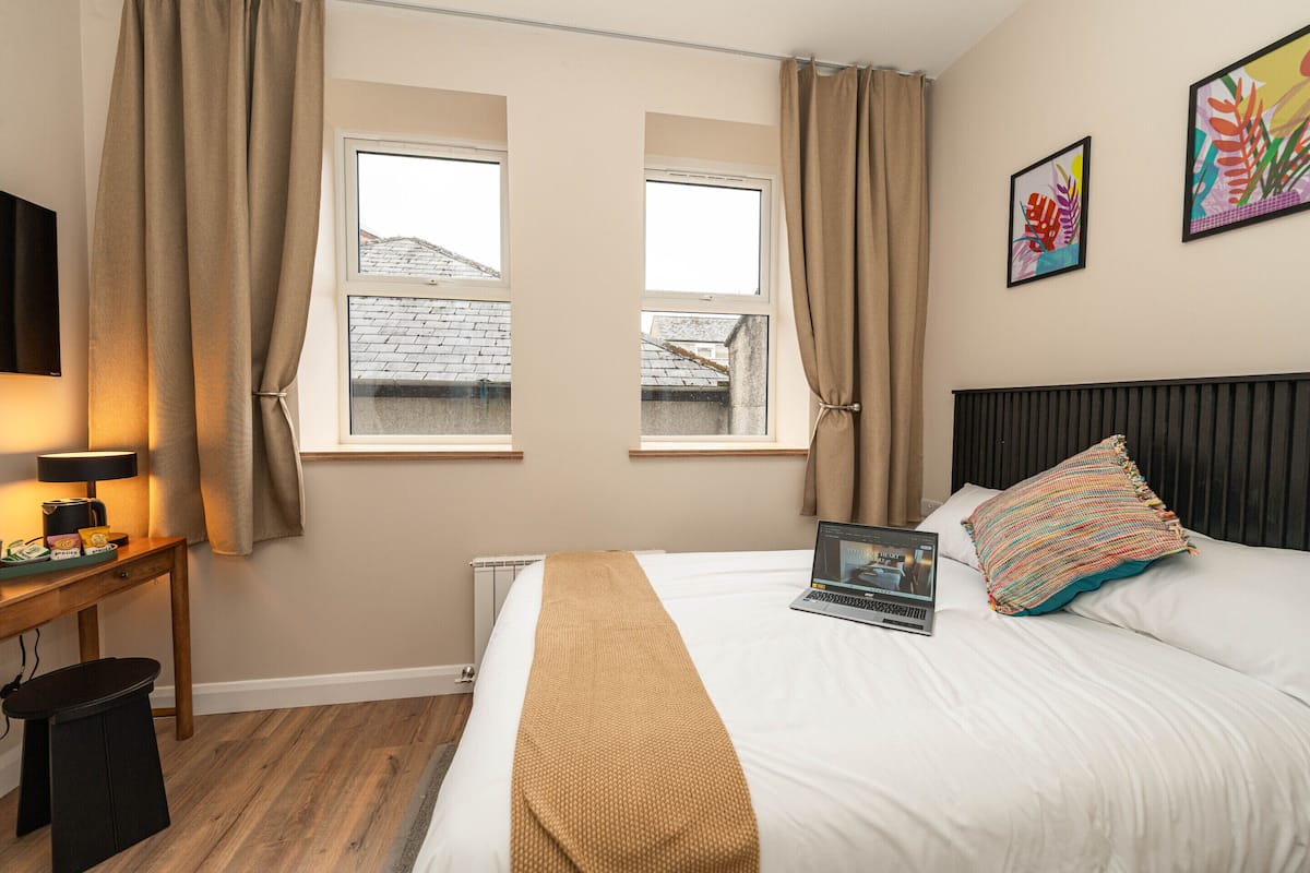A hotel room with a bed, a desk with a laptop, a radiator, and two windows with curtains.