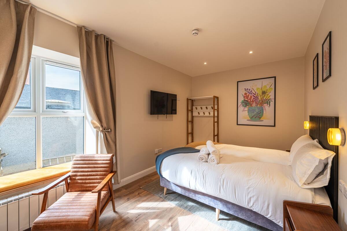 A bedroom with a bed, a chair, a window, a painting, and a television.