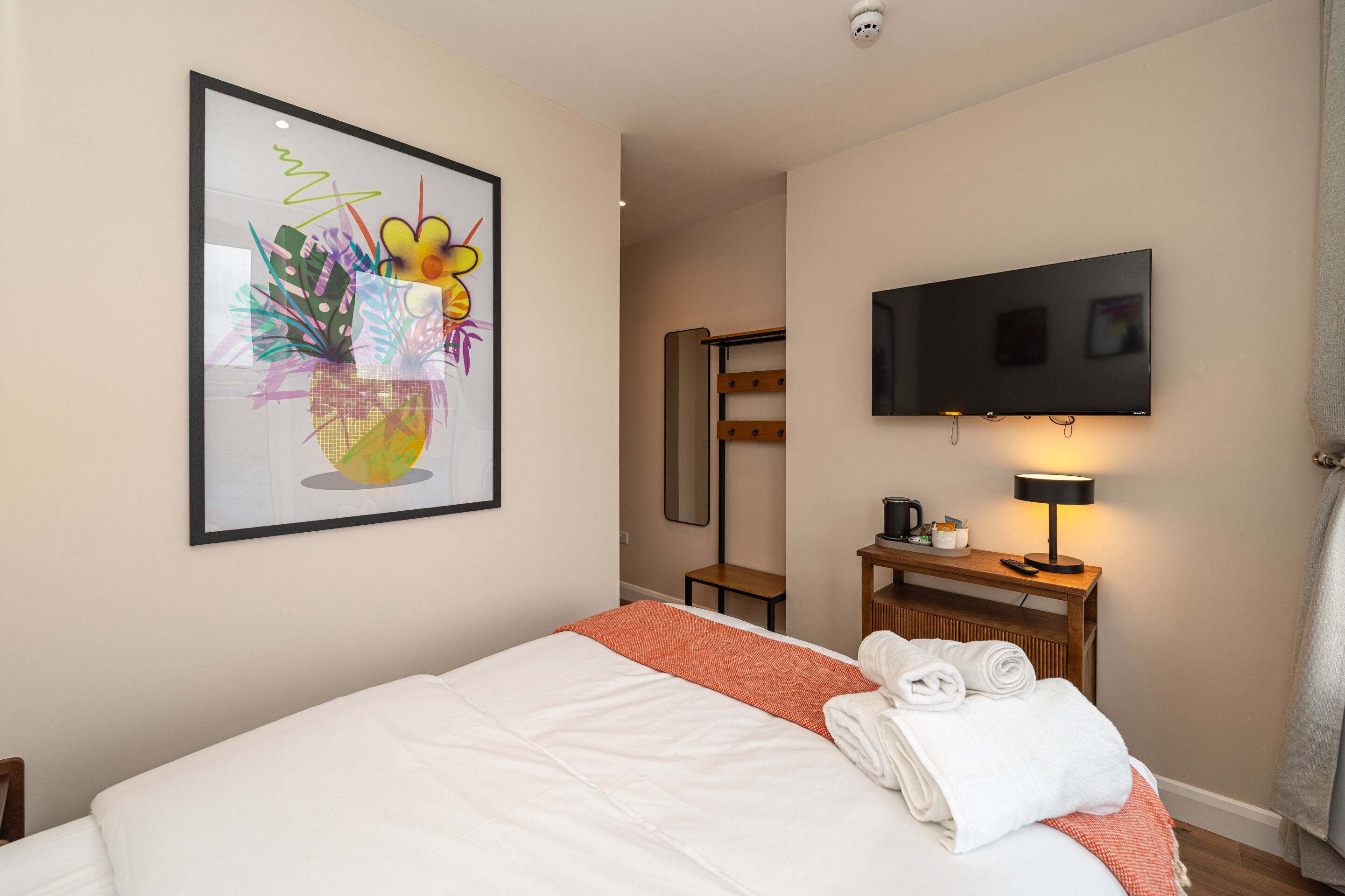 A hotel room with a bed, a television mounted on the wall, a framed artwork above the bed, and a small table with a lamp and a coffee maker.