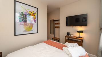 Une chambre d’hôtel avec un lit, un téléviseur fixé au mur, une œuvre d’art encadrée au-dessus du lit, et une petite table avec une lampe et une machine à café.