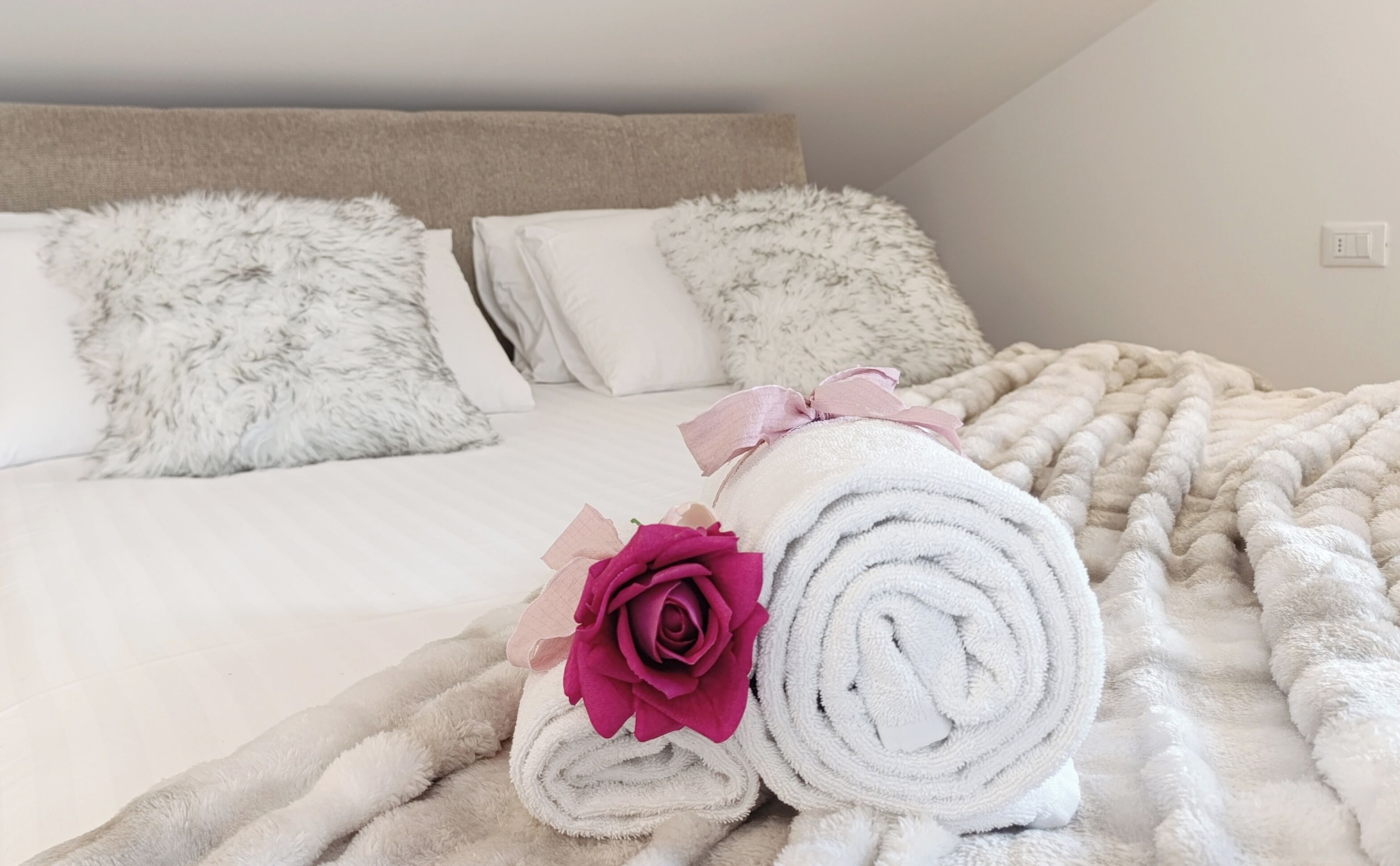Una cama impecablemente tendida con almohadas mullidas, una toalla blanca enrollada con una rosa rosa, y un edredón de color claro.