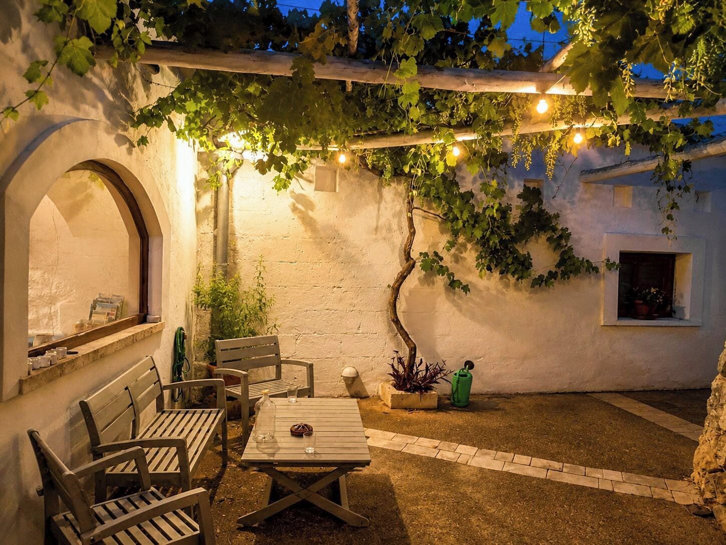 A cozy outdoor seating area with a table and benches, surrounded by plants and a pergola with string lights.