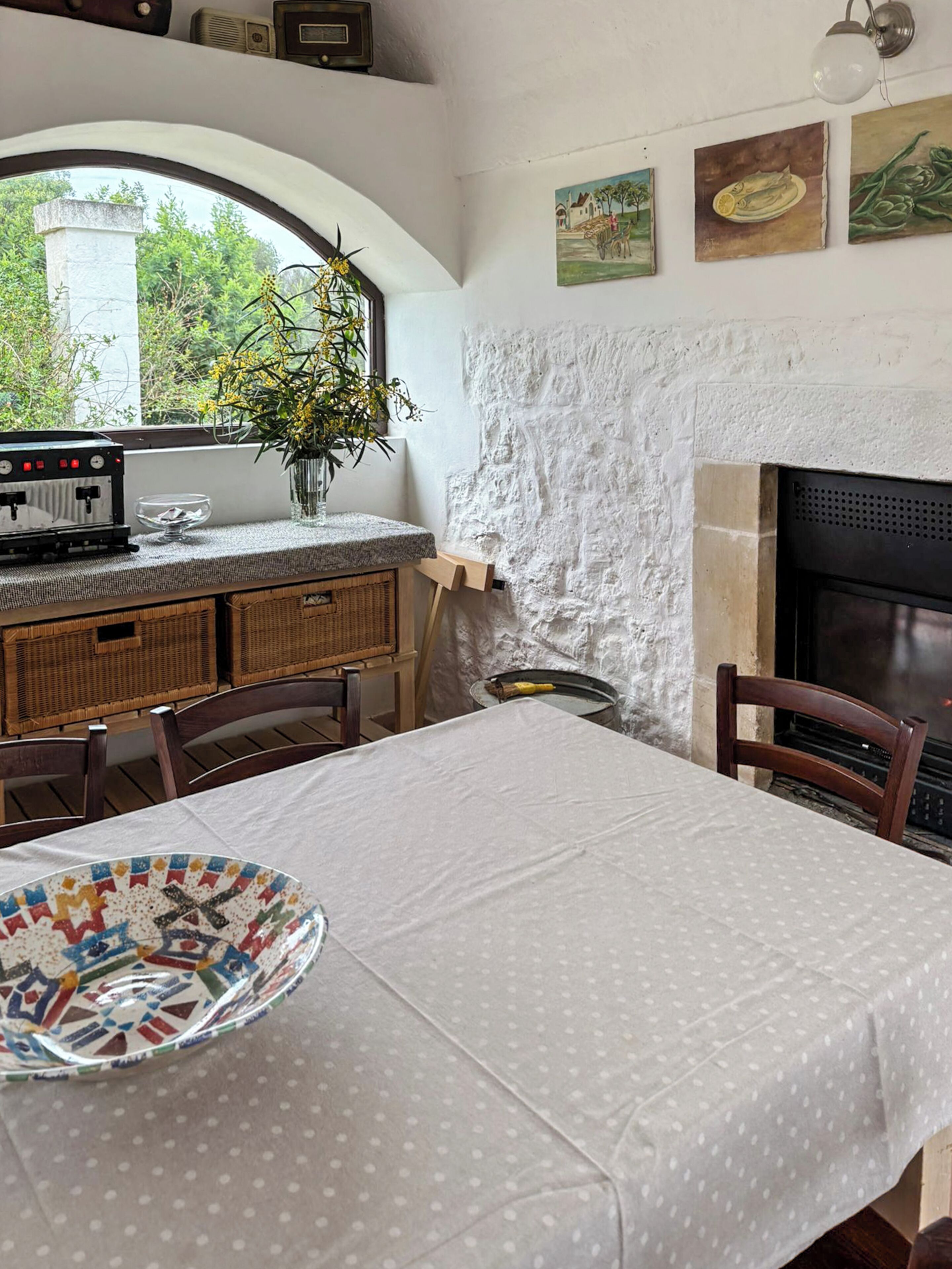 A dining area with a table set for two, a fireplace, a window with a view of greenery, and a wall with artwork.
