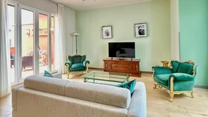 Ein Wohnzimmer mit einer Couch, zwei Sesseln, einem Glastisch, einem Fernseher und einer Glasschiebetür zu einem Außenbereich.