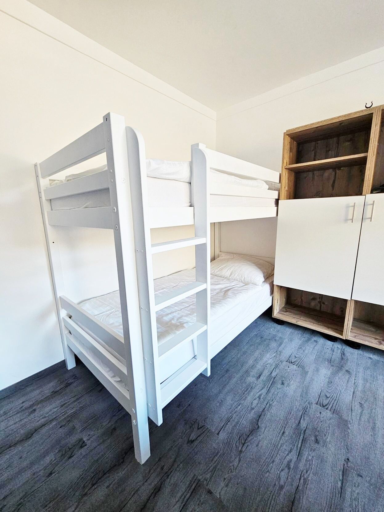 Ein Etagenbett mit Holzrahmen und weißer Bettwäsche, ein Holzschrank mit offenen Regalen und ein grauer Holzboden.