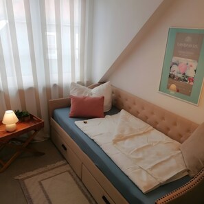 Une chambre à coucher avec un lit, une table de chevet avec une lampe, un tableau encadré au mur et une fenêtre avec des rideaux légers.