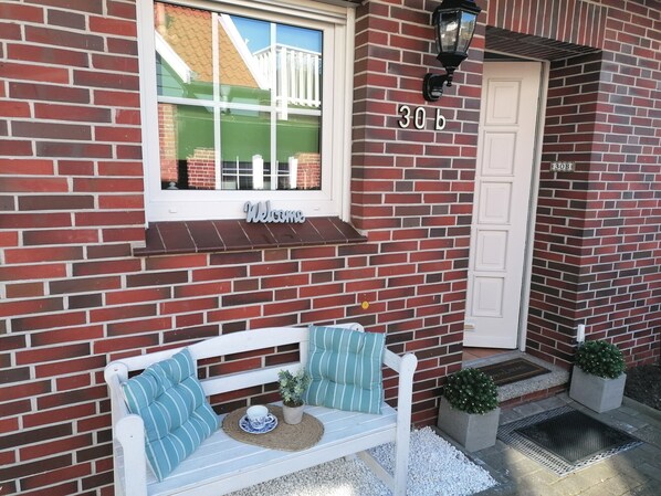 Un bâtiment en briques avec une porte blanche, une pancarte indiquant « Bienvenue » et un banc à l’extérieur.