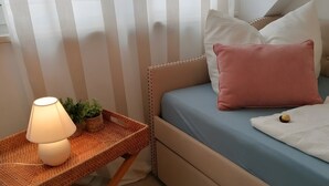 Ein Schlafzimmer mit einem Bett, einem Nachttisch mit Lampe, einer kleinen Pflanze und einem Fenster mit Vorhängen.