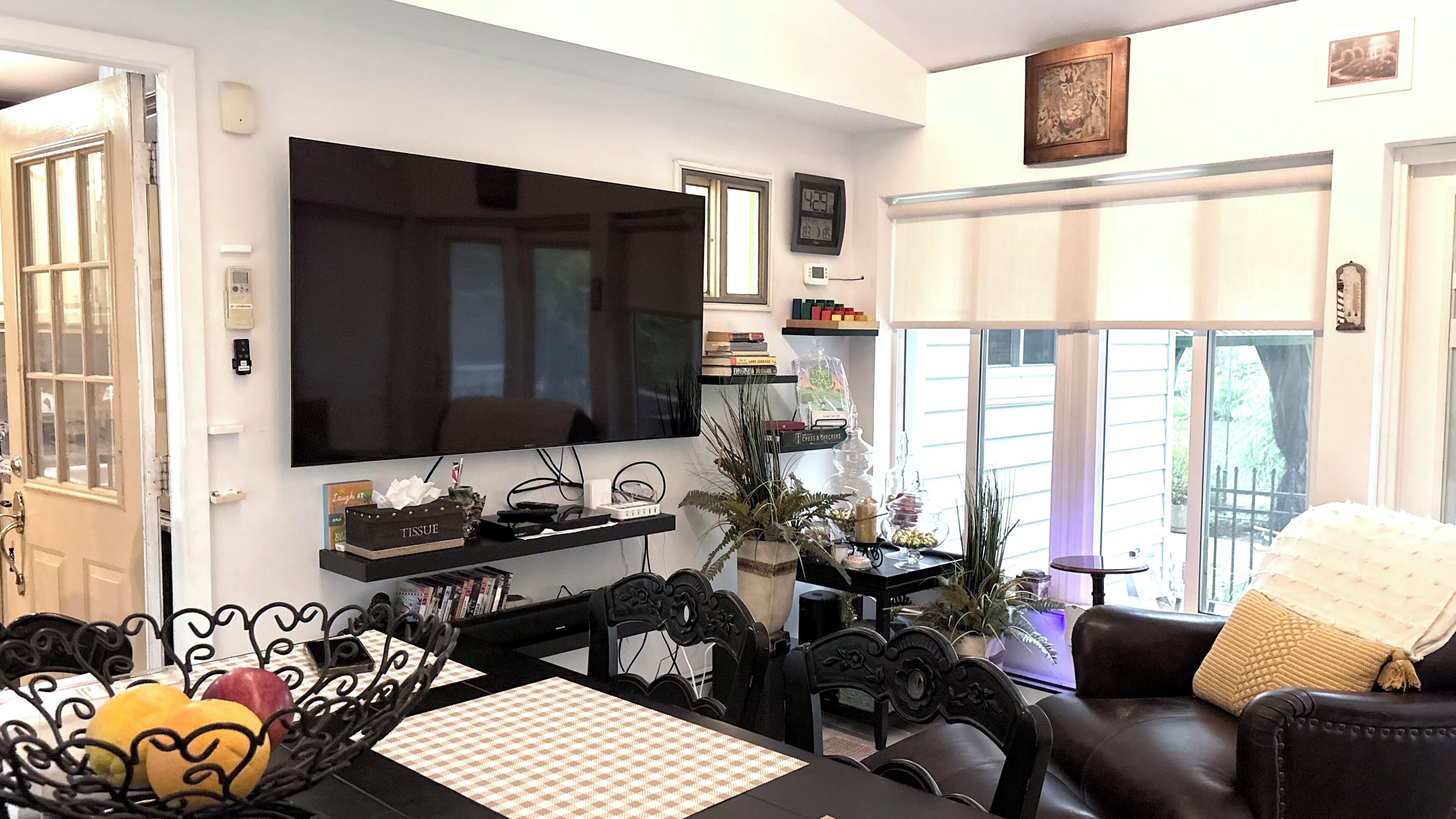 A living room with a flat-screen TV, a dining table with a placemat, a sofa with a throw blanket, and a window with blinds.
