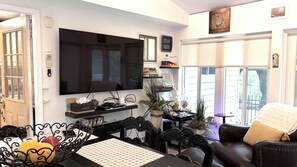 A living room with a flat-screen TV, a dining table with a placemat, a sofa with a throw blanket, and a window with blinds. - Peaceful 3BR Garden Cottage Long Island Escape (Coram)
