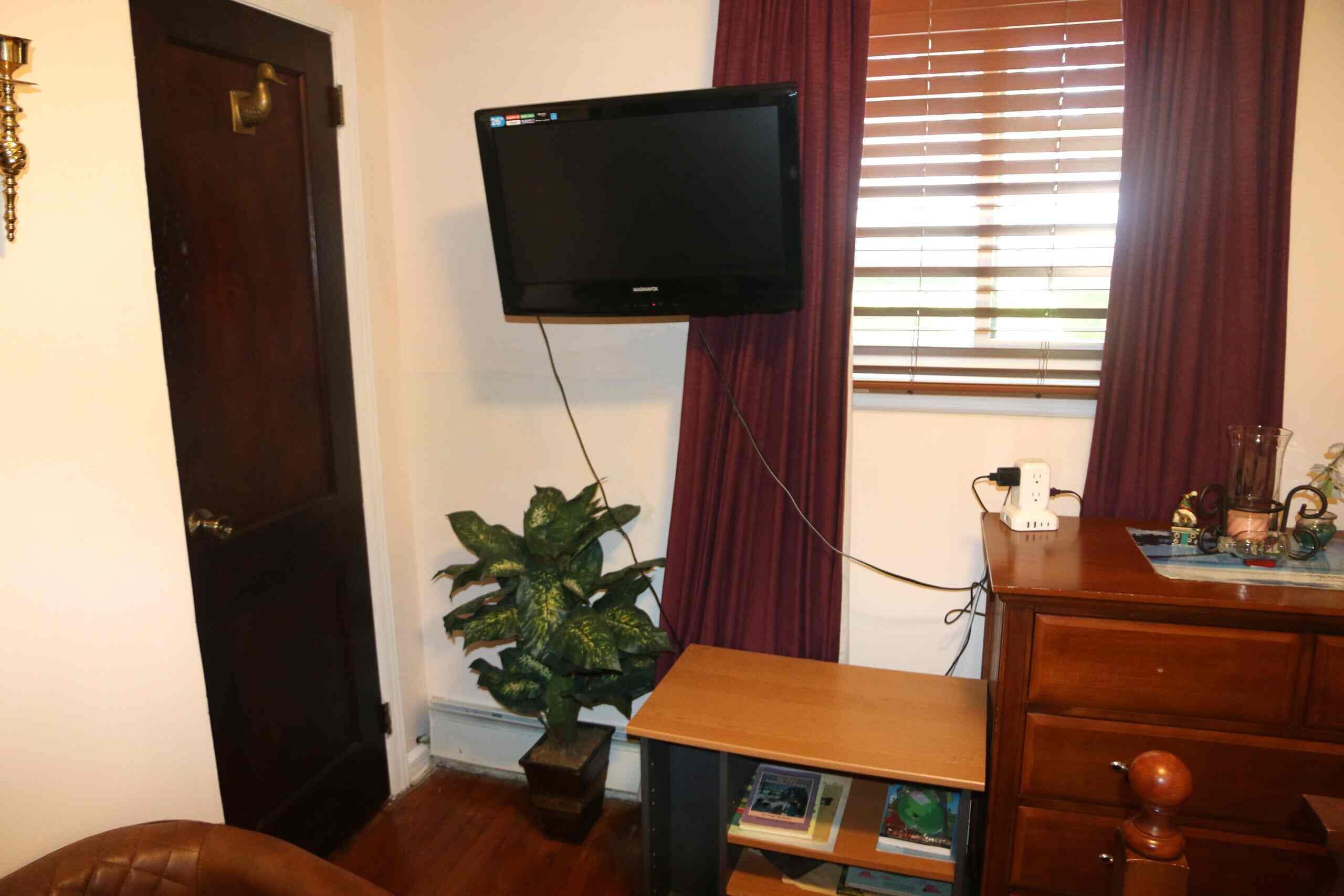 Ein Zimmer mit einem an der Wand befestigten Fernseher, einem Holzschrank, einer Pflanze und einem Fenster mit Vorhängen.