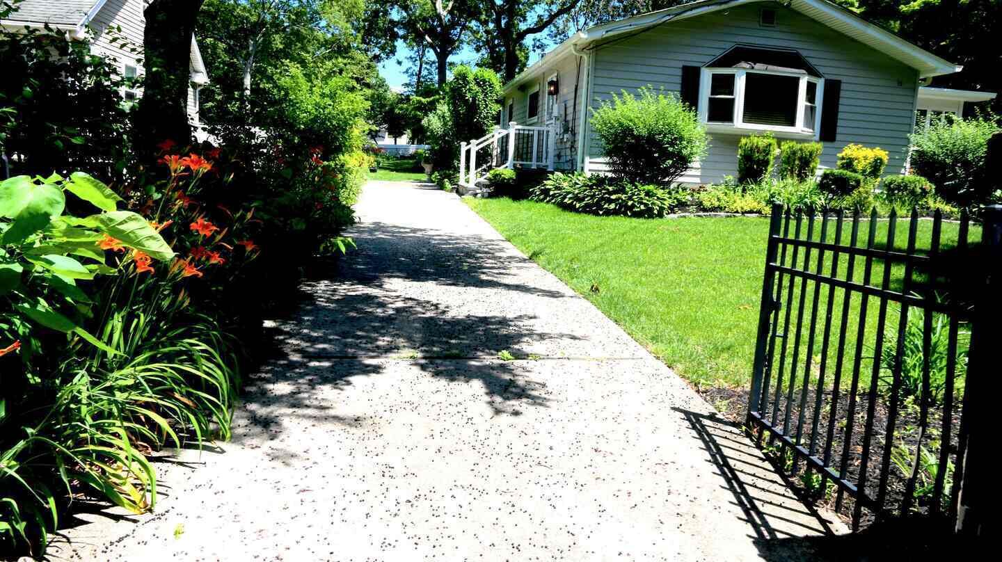 A residential driveway leading to a house with a white exterior, surrounded by greenery and a black metal fence.