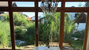 A dining area with a white table, four chairs, and a chandelier. Large windows offer a view of greenery and a wooden structure outside. - Wonderfully private vacation home near the lake on Lake Chiemsee with large garden & terrace (Gstadt am Chiemsee)