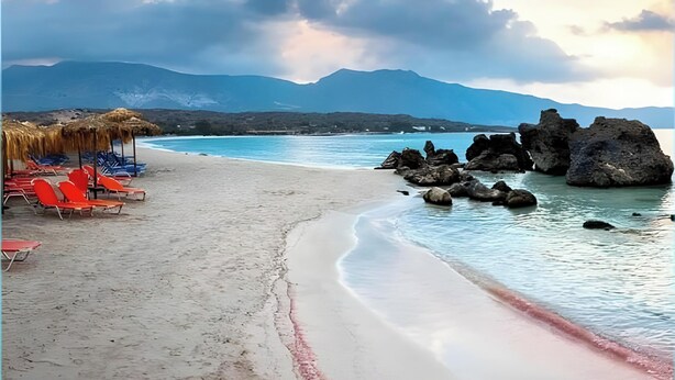 Une plage avec un ciel dégagé, une mer calme et des montagnes en arrière-plan.