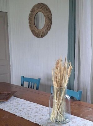 Una mesa de comedor con mantel floral, un florero de vidrio transparente con plantas secas, una silla azul y un espejo redondo en la pared.