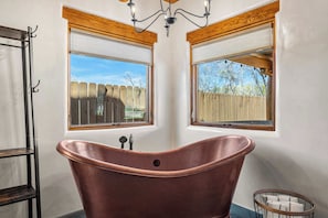 A bathroom with a freestanding copper bathtub, two windows with wooden frames, and a chandelier.
