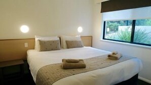 A hotel room with a bed, pillows, a window, and a bedside table. - Margaret River Beach Apartments (Gnarabup)
