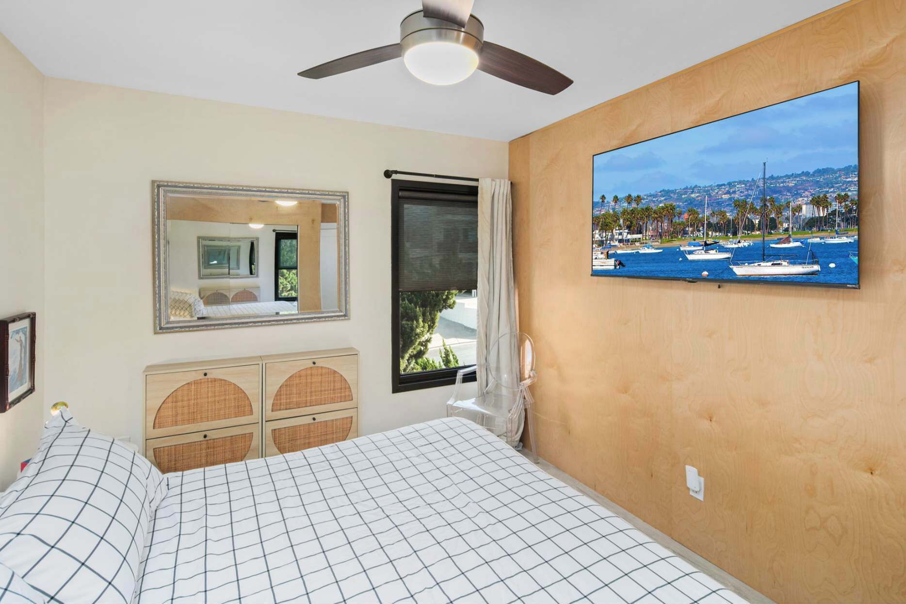 A bedroom with a bed, a ceiling fan, a large window with a view of boats, a mirror, and a wooden headboard.