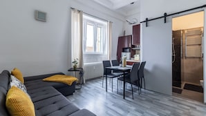 A modern living room with a sofa, dining table, and chairs, a kitchen area with cabinets and a microwave, and a sliding door leading to a shower area. - Panoramic Top-Floor Downtown Apartment with Citadel & Liberty Statue View (Budapest)