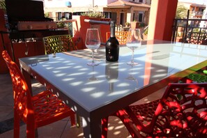 Ein Tisch mit Glasplatte, zwei Weingläsern und einer Flasche, umgeben von roten Stühlen und einem Grill im Hintergrund.