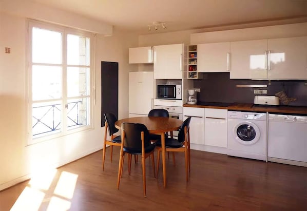 A modern kitchen with white cabinets, a dining table with chairs, a washing machine, and a large window. - Le skipper - Les Gîtes de la Côte d'Amour (LE CROISIC)