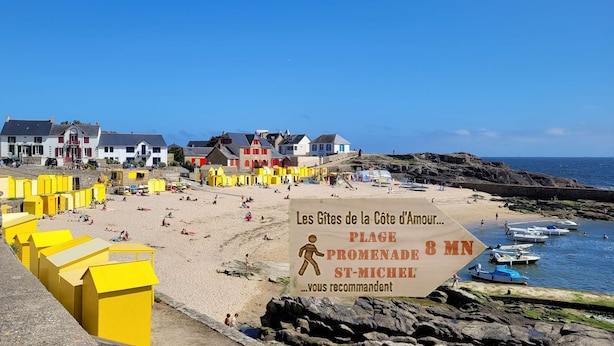 En strand med gula strandhytter, en skylt som visar att det är åtta minuters promenad längs strandpromenaden till Saint-Michel, och båtar i vattnet.