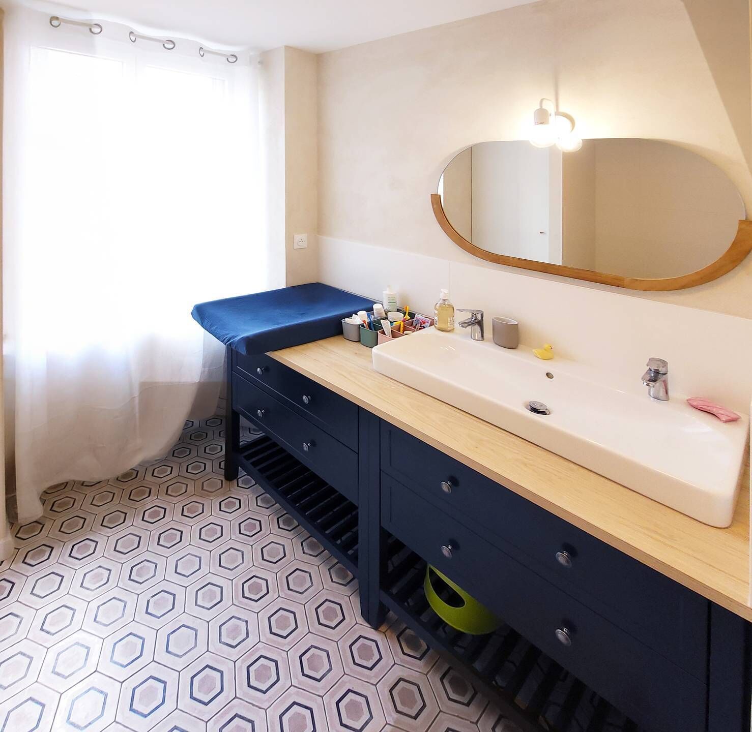 A bathroom with a large mirror, a sink, a blue bench, and a hexagonal patterned floor.