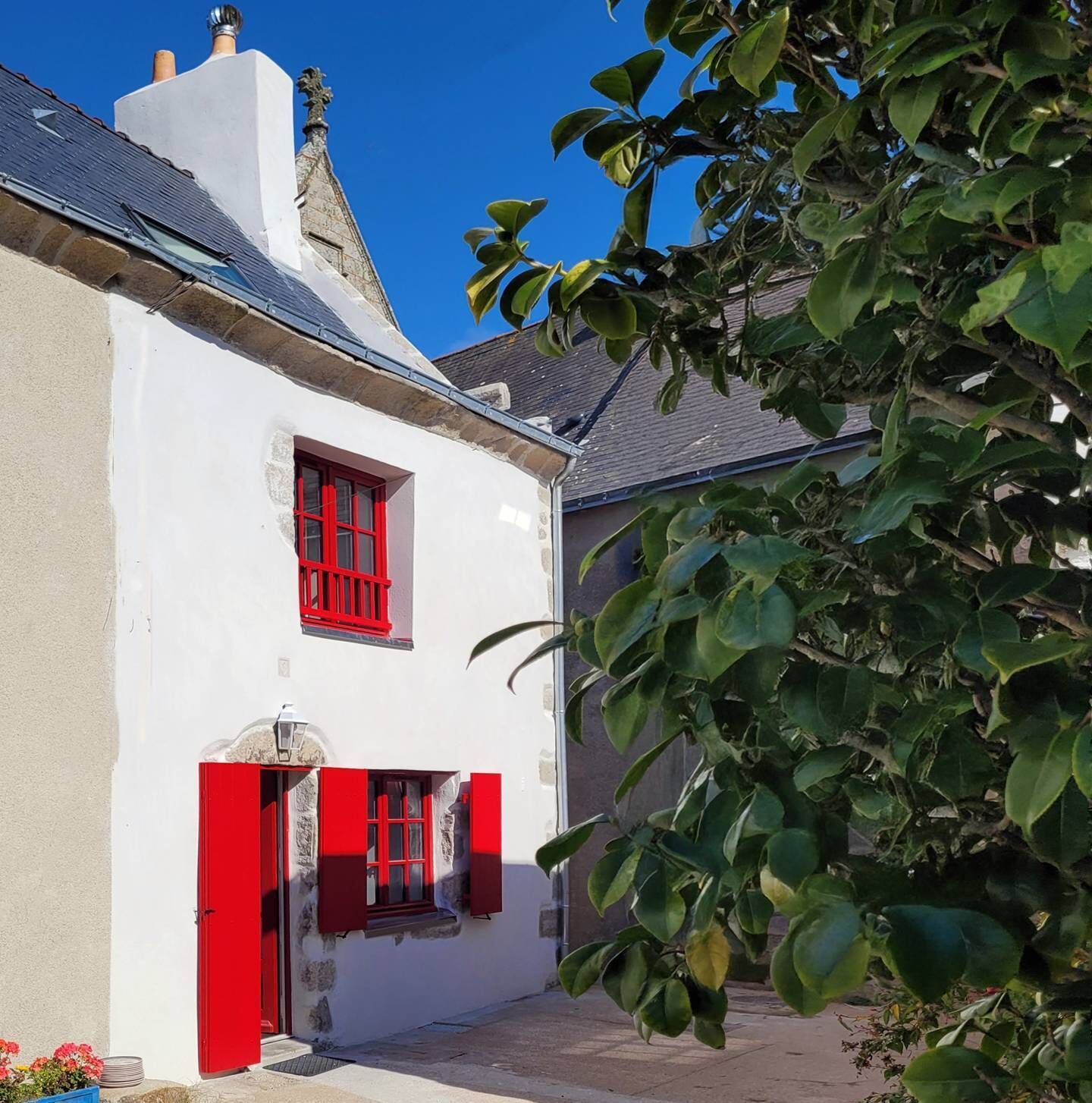 Un bâtiment blanc avec des volets rouges et une porte, un ciel dégagé et un arbre au premier plan.