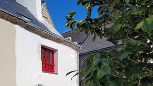 A white building with red shutters and a door, a clear sky, and a tree in the foreground. - Le clos du prieuré - Les Gîtes de la Côte d'Amour (BATZ SUR MER)
