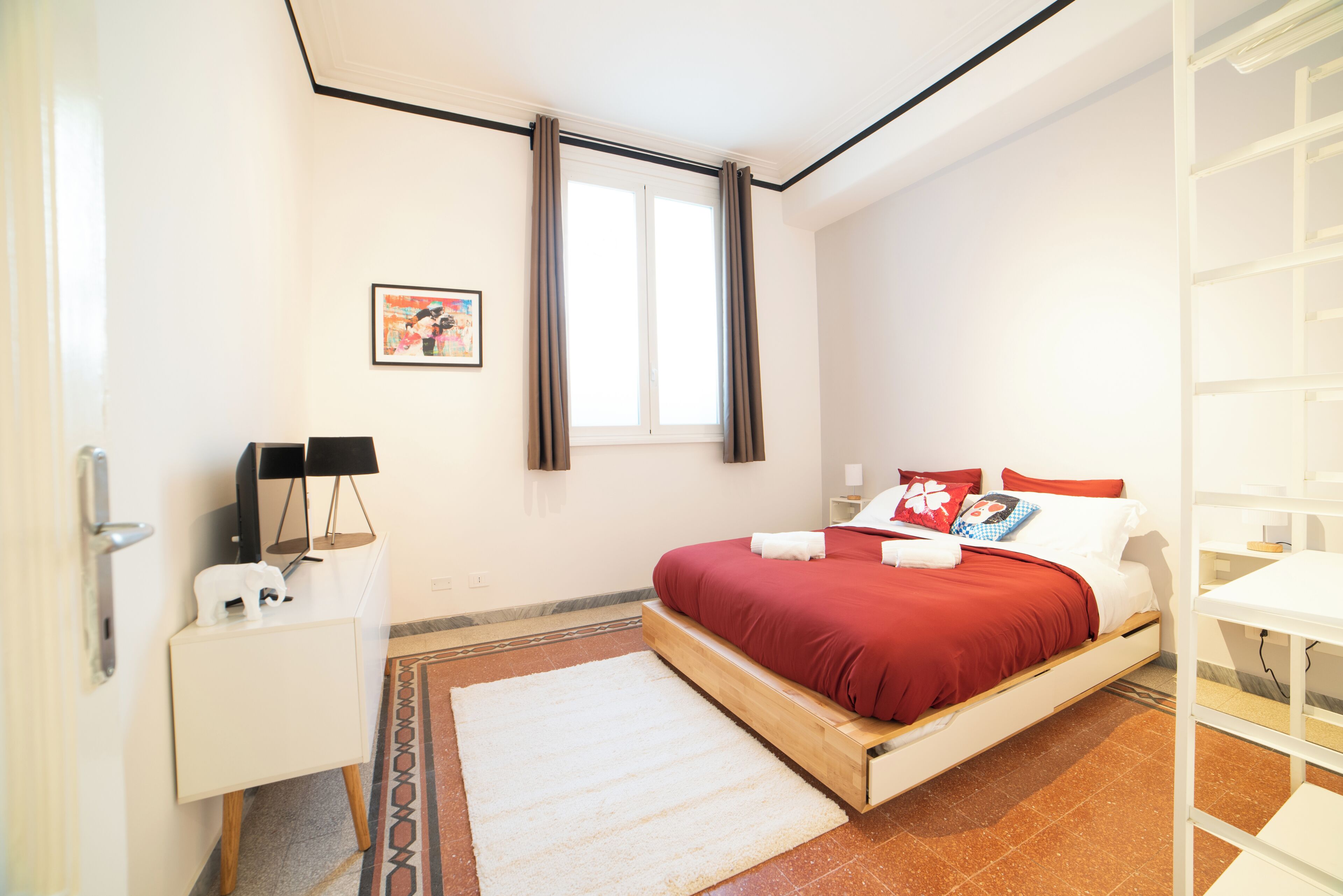 A bedroom with a bed, a bedside table, a nightstand, a window with curtains, and a wall-mounted picture.