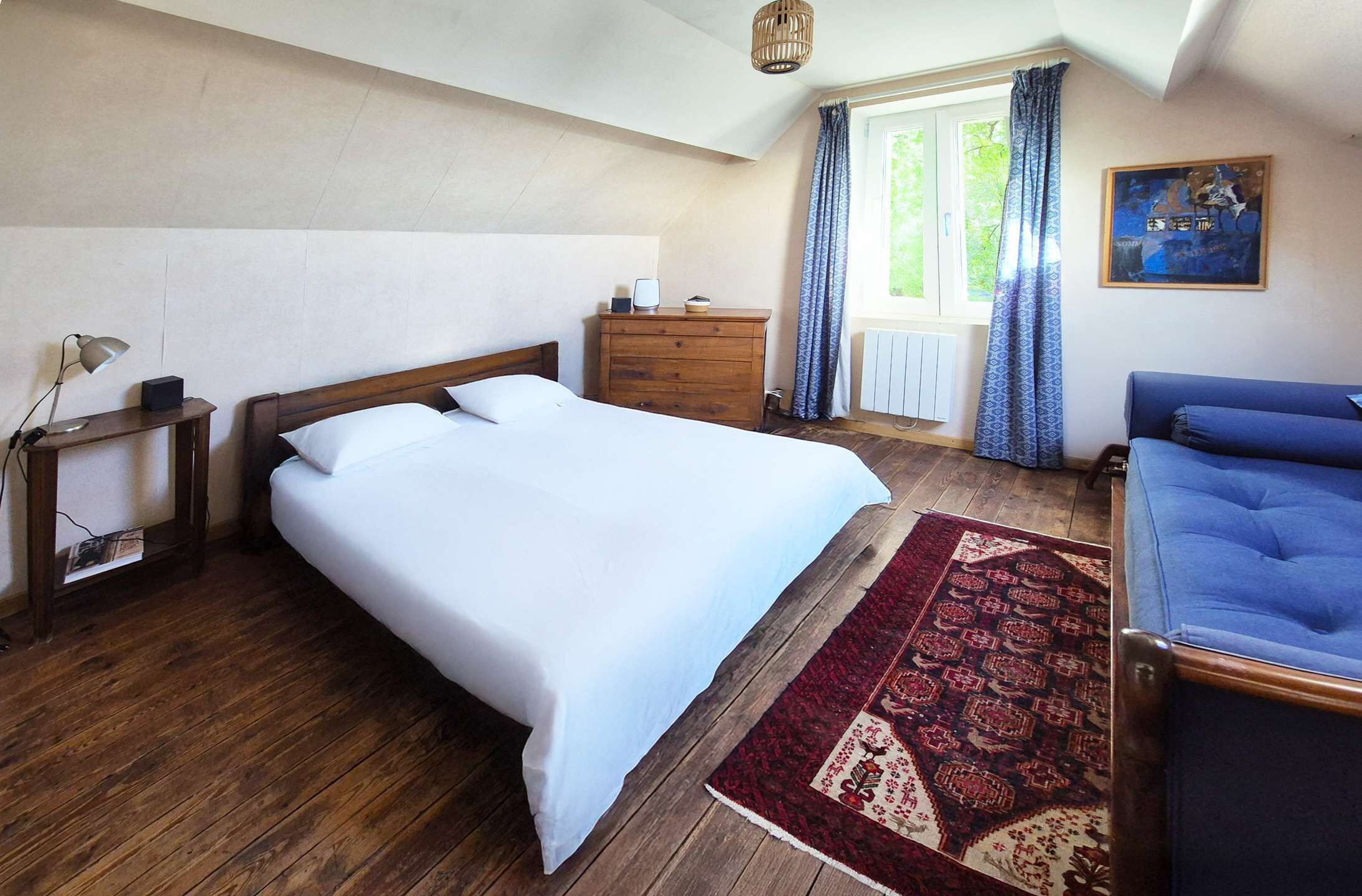 A bedroom with a large bed, a wooden dresser, a blue sofa, a patterned rug, and a painting on the wall.