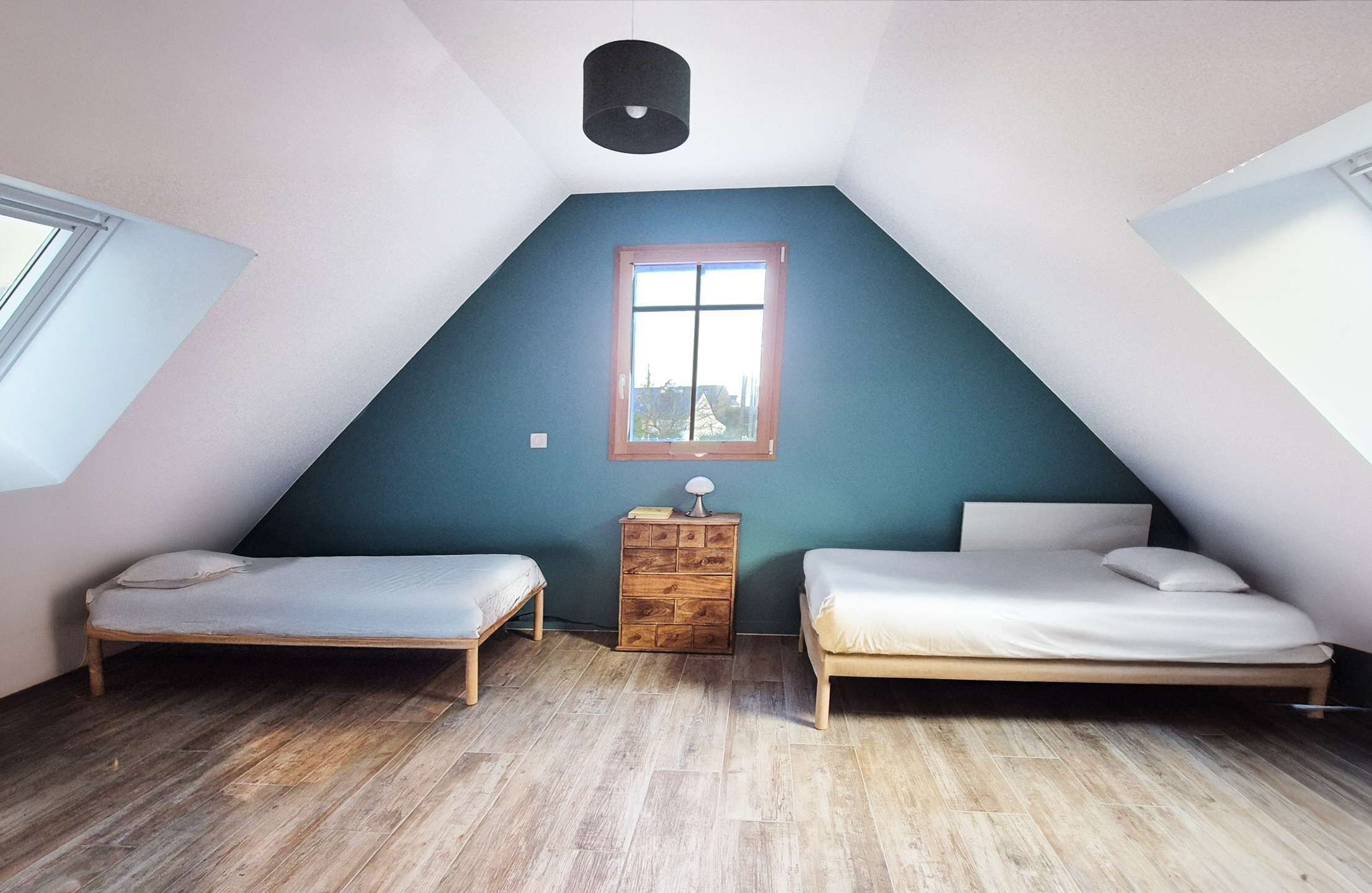 Ein Zimmer mit zwei Einzelbetten, einem Holzschrank, einem Fenster und einer schrägen Decke.