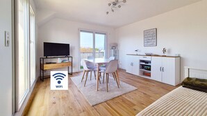 A modern living room with a TV, dining table, chairs, and a kitchen area with cabinets. - Le nid de port-lin - Les Gîtes de la Côte d'Amour (LE CROISIC)