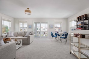 A modern living room with a sofa, a dining table with chairs, a bookshelf, and a balcony with large windows. - Harborside | Golf | Pool | Marina Views (St Joseph)