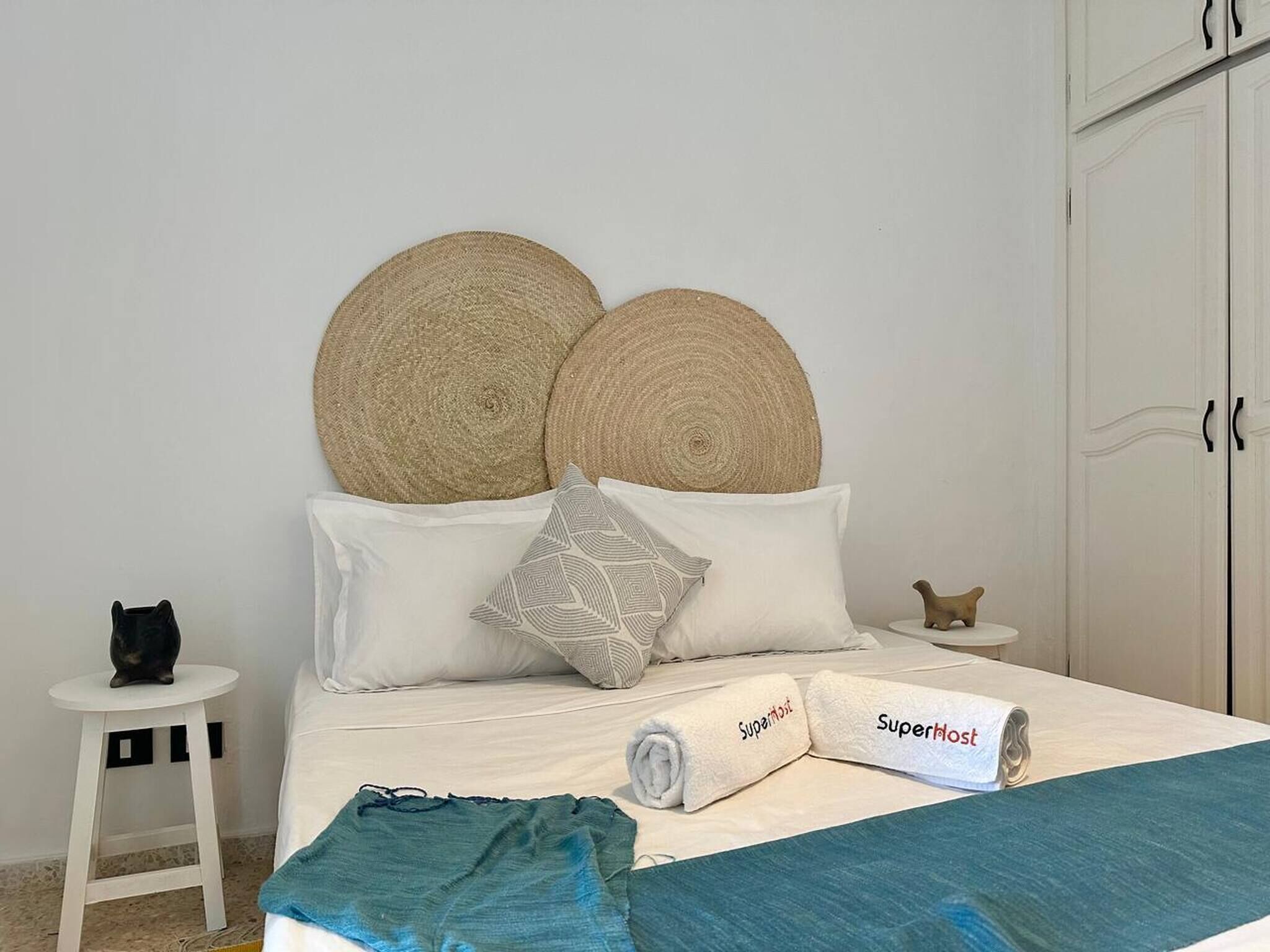 Un letto con testiera, cuscini, un asciugamano arrotolato, un tavolino con un gatto e un armadio.