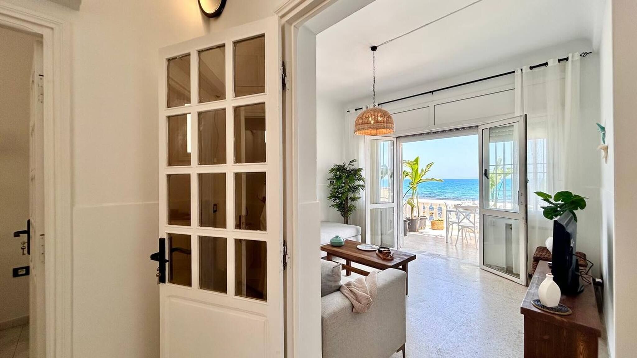 A hallway leading to a balcony with a view of the ocean, a dining area with a table and chairs, and a large window with multiple panes.