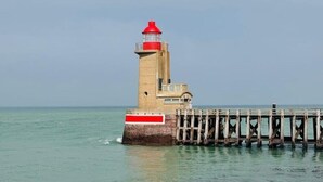 Un phare avec un sommet rouge, une base en béton et une jetée en bois qui s’avance dans l’eau.
