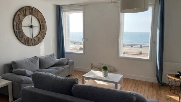 Un salon moderne avec un canapé gris, une horloge ronde en bois, une table basse blanche et une vue sur la mer grâce à de grandes fenêtres.