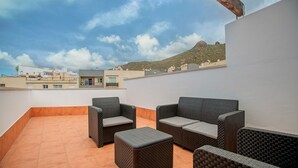 Una terraza en la azotea con mobiliario de mimbre, piso de baldosas y vistas a edificios y una colina.