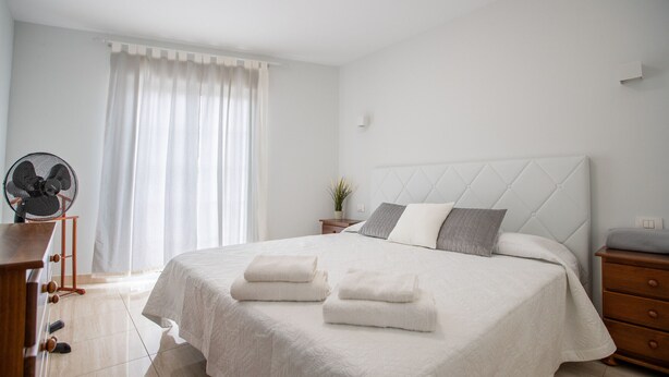 Un dormitorio ordenado con cama, mesitas de noche, un ventilador y una ventana con cortinas.