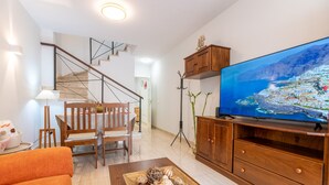 Una sala de estar con una escalera de madera, una mesa de comedor con sillas, un televisor sobre un mueble de madera y una bandeja con objetos decorativos sobre una mesa auxiliar.