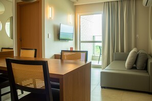 Una sala moderna con una mesa de comedor de madera, sillas de mimbre, un sofá con cojines, un televisor de pantalla plana y un ventanal corredero que da a un balcón.