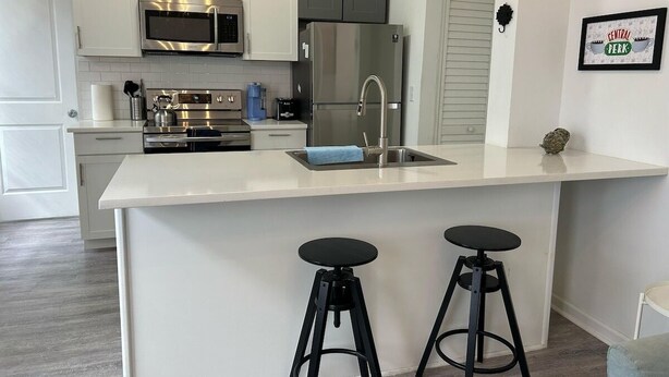 A modern kitchen with white cabinets, a stainless steel refrigerator, a double oven, and two black stools. - Trendy Bungalow near Armature and Downtown Tampa (Tampa)