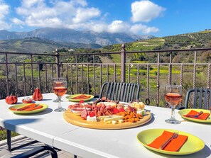 A table set outdoors with a scenic mountain view, a variety of cheeses, meats, and fruits, and two glasses of red wine. - MUSES MOUNTAIN VIEW (Gortyna)