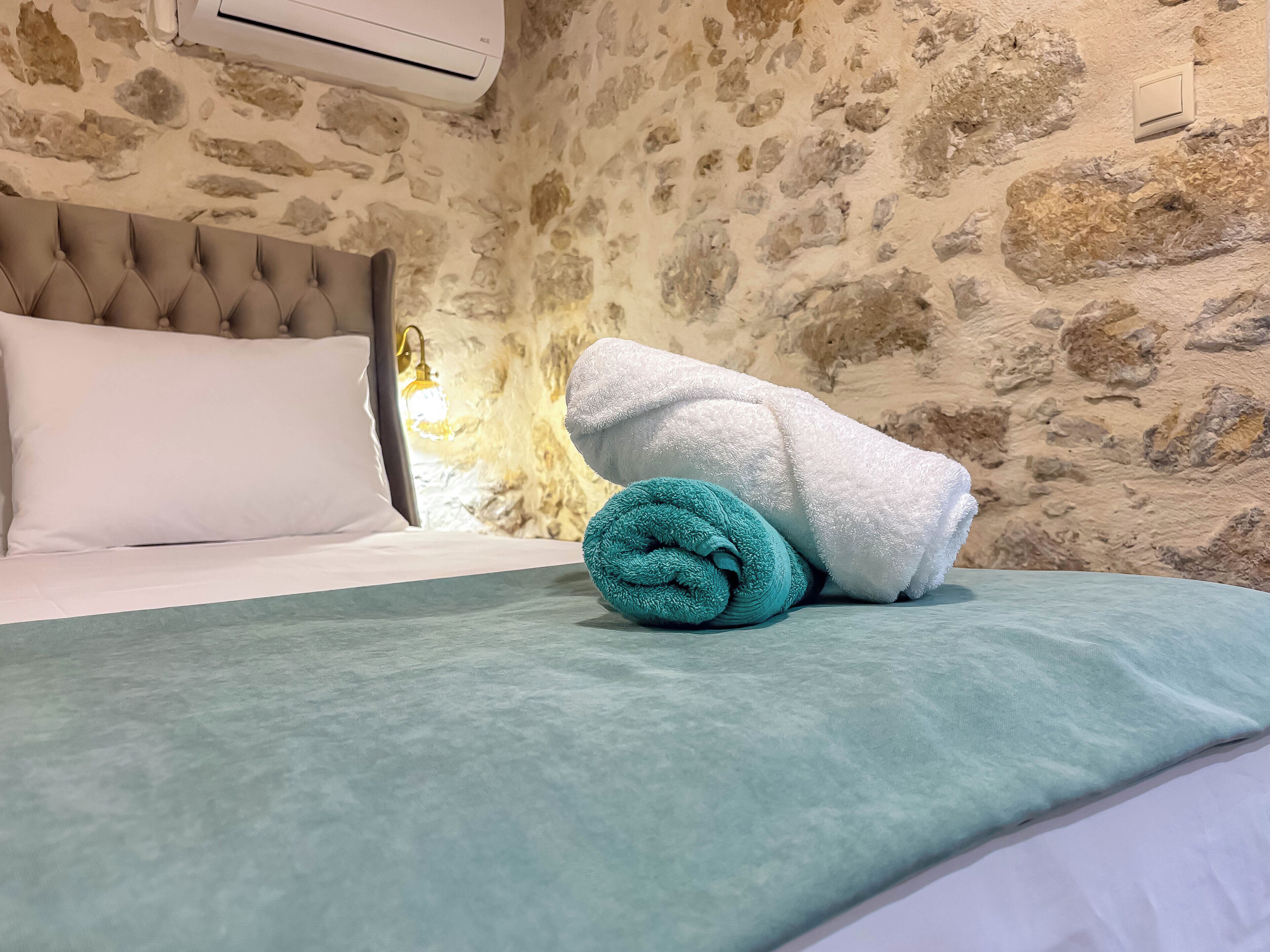 Un lit avec un oreiller blanc, une serviette turquoise et une couvre-lit vert clair, dans une pièce aux murs en pierre et dotée d’un climatiseur blanc.