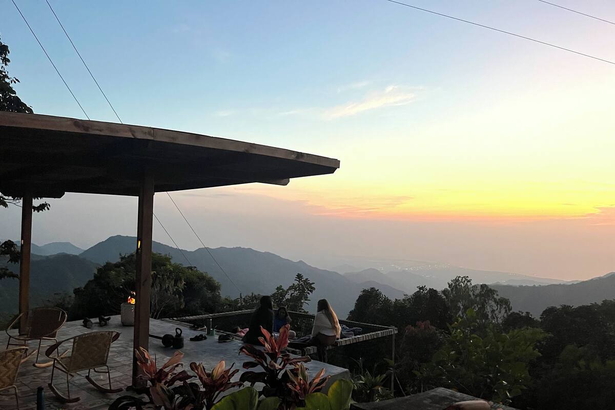 A terrace with seating and potted plants, overlooking a mountainous landscape during sunset.