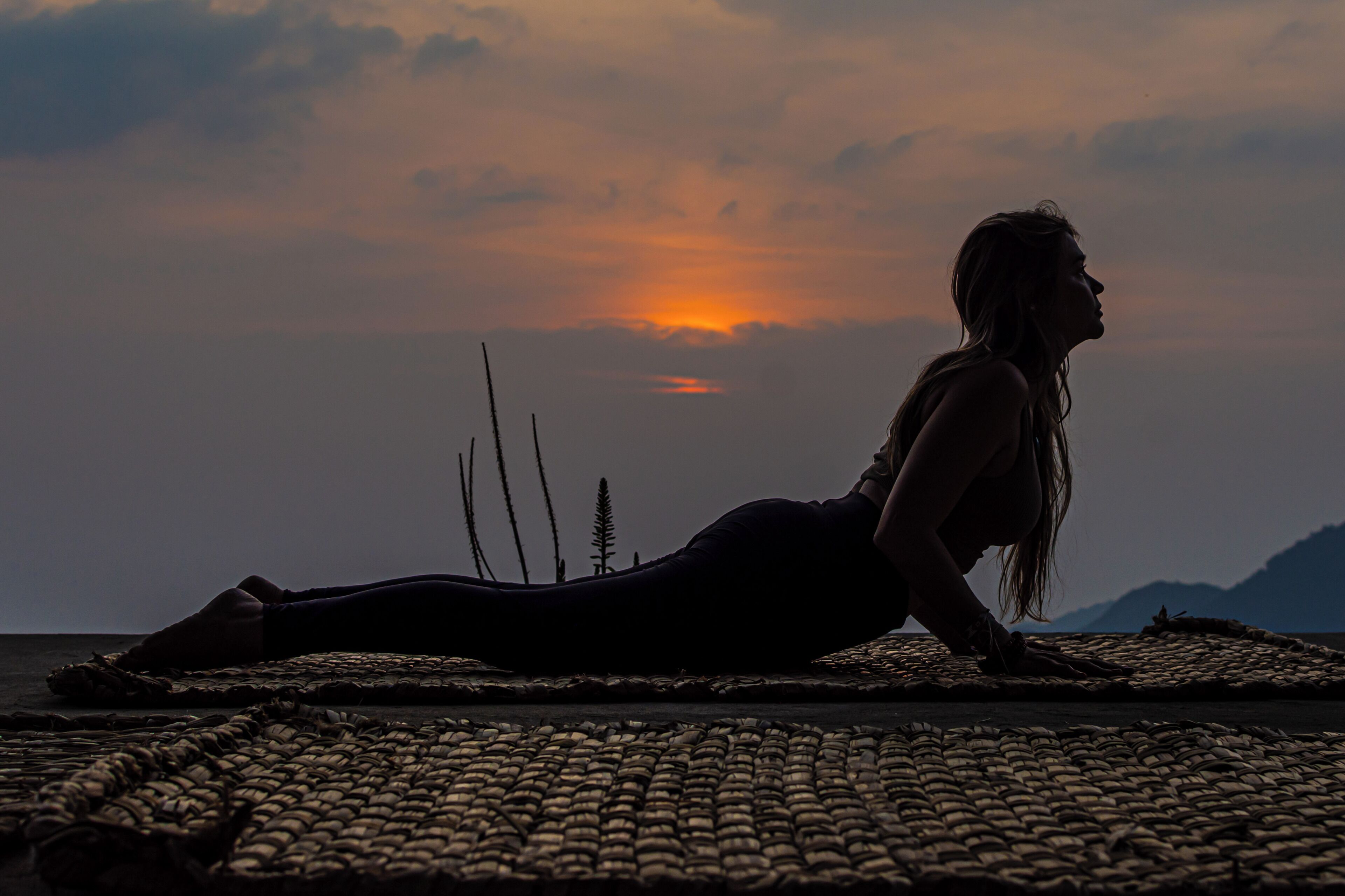 Eine Person in einer Yoga-Pose auf einer gewebten Matte vor einem Sonnenuntergang.